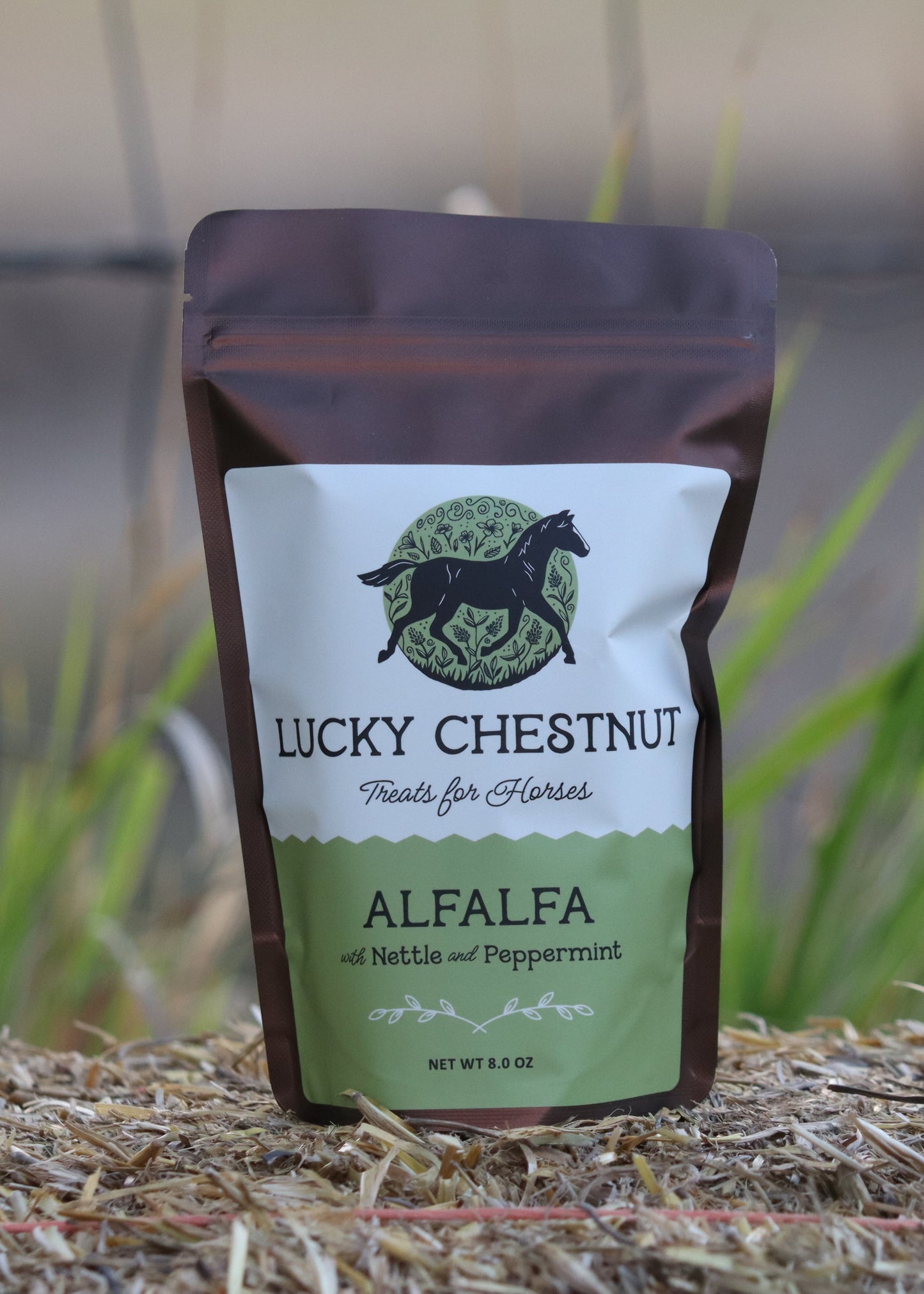 Lucky Chestnut Alfalfa horse treats pouch with nettle and peppermint, displayed upright on straw with green plants in the background.