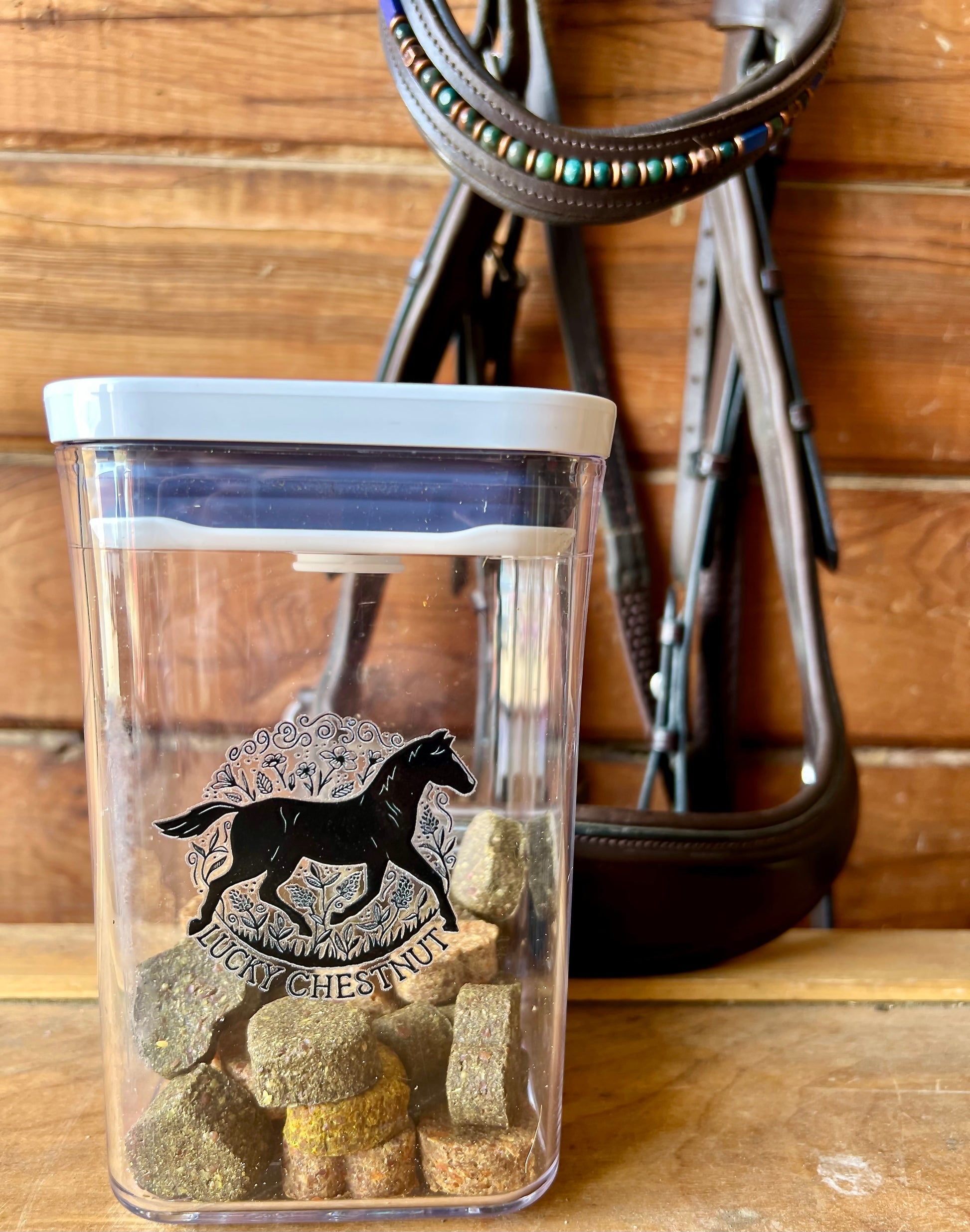 Front view of Lucky Chestnut clear horse treat container filled with treats, set on a wood surface with a bridle in the background.